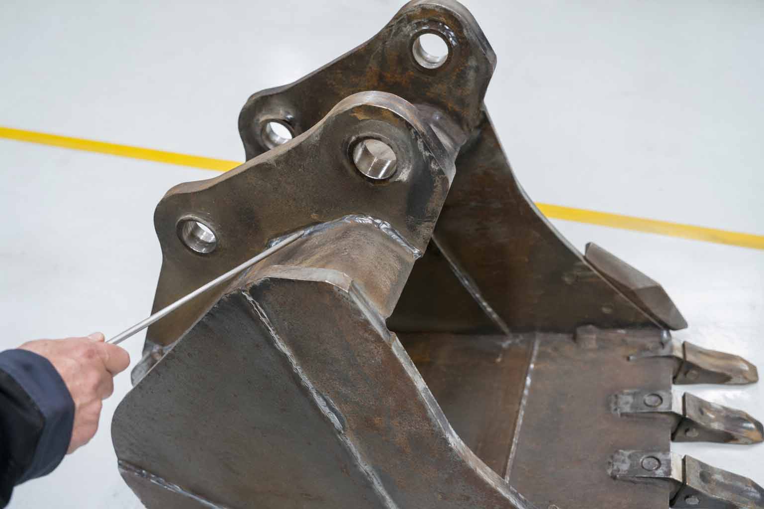 Close-up of excavator bucket showing material thickness, weld seams, and wear areas in a factory setting